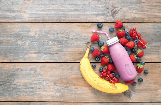 Berry smoothie in a bottle with ingredients on top of a wooden background with space 