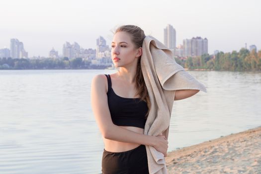 Young pretty slim fitness sporty woman rests and wipe herself with towel during training workout exercises outdoor at river coast in the morning