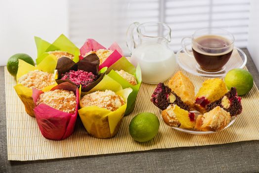 Color muffins in multicolored paper, with milk, a cup of coffee and limes on a mat from a bamboo and a cloth from rough fabric