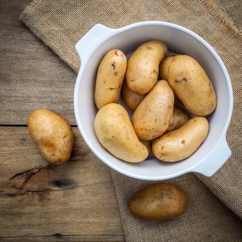 Composition of fresh organic potatoes in white ceramic bowl on hemp sake and   rustic wooden table. Preparation for cooking.