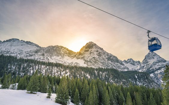 Winter scenery with the sun hiding behind the Alps mountains, covered by fir forests, and a cable car crossing them. Image captured in Ehrwald, Austria.