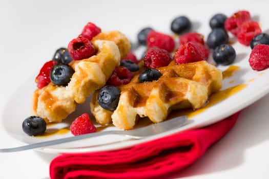 Close-up of waffles with berries and maple syrup on a white plate over a red napkin