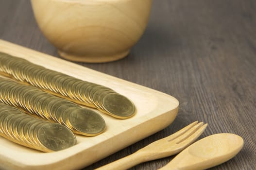 Close up stack of gold coins in wooden tray have blur spoon with fork and cup on wood background.