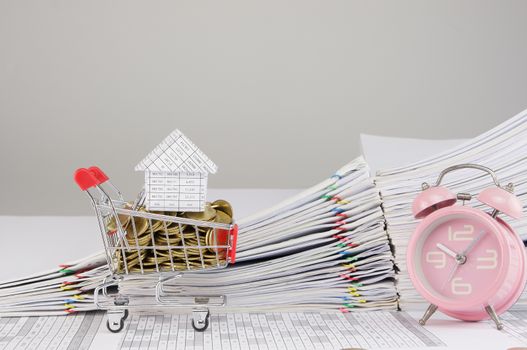 House on shopping cart with gold coins and old pink alarm clock on finance account have blur pile overload document of report and receipt with colorful paperclip as background.