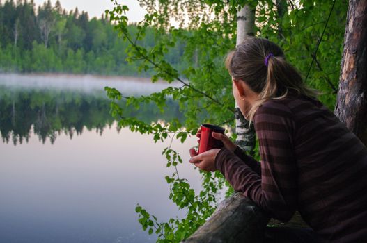 a beautiful young girl drinking cup of coffee or tea. Portrait of attractive woman thoughtfully looks out over lake with water reflection and holding cup of tea to warm up.