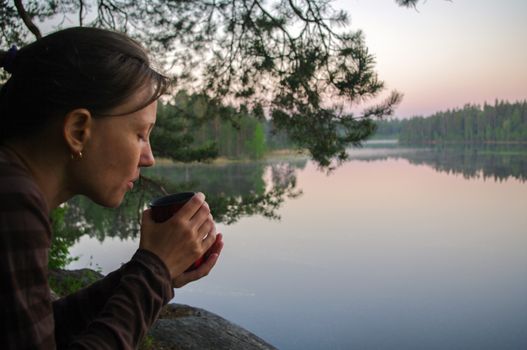 a beautiful young girl drinking cup of coffee or tea. Portrait of attractive woman thoughtfully looks out over lake with water reflection and holding cup of tea to warm up.