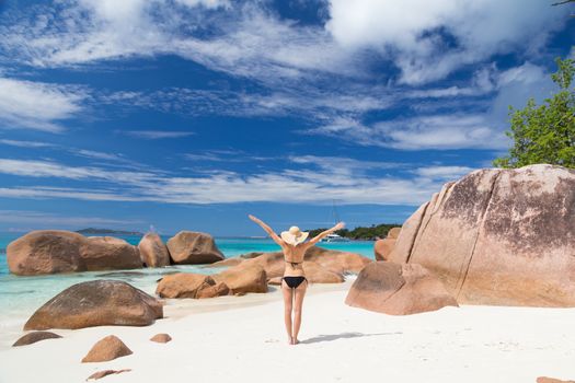 Woman arms rised, wearing black bikini and beach hat, enjoying amazing view on Anse Lazio beach on Praslin Island, Seychelles. Summer vacations on picture perfect tropical beach concept.