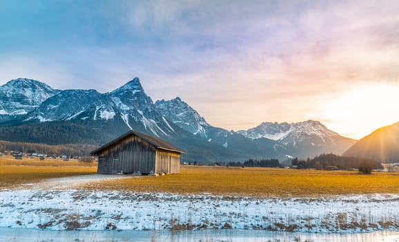 Winter alpine landscape with an aged wooden barn, the Alps mountains, a frozen river at sundown. Picture captured in Austria.