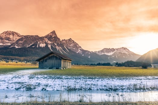 Winter landscape with an old wooden barn on a snowy field, a frozen river, the Austrian Alps mountains and a December sun setting down.