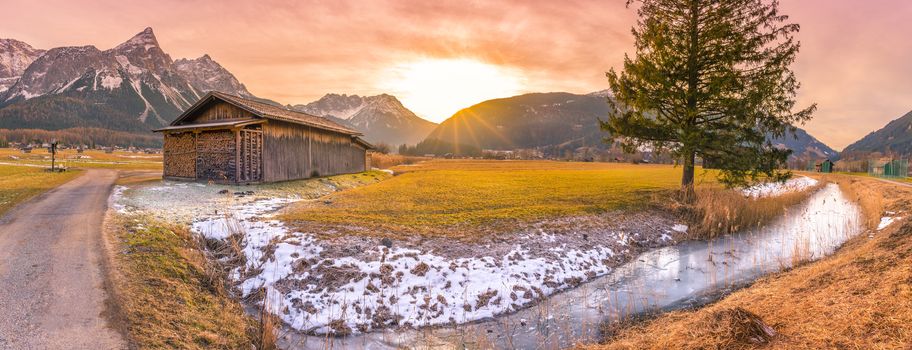 Idyllic winter scenery in a small Austrian village, with a wooden barn, a frozen river and the Alps mountains,  at dusk.