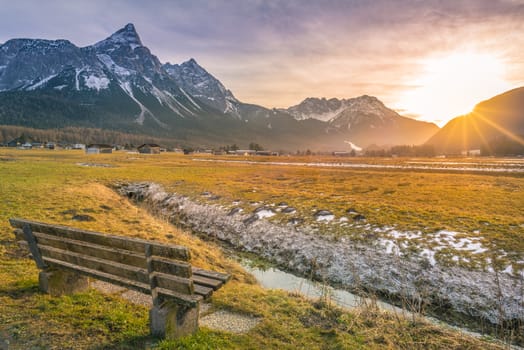 Charming winter scenery with a wooden bench on the side of a small river with a view towards the Austrian Alps, at sunset.