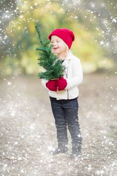 Baby Girl In Red Mittens and Cap Holding Small Christmas Tree Outdoors with Snow Effect.