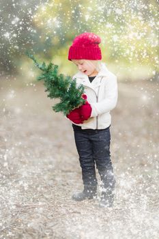 Baby Girl In Red Mittens and Cap Holding Small Christmas Tree Outdoors with Snow Effect.