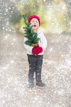 Baby Girl In Red Mittens and Cap Holding Small Christmas Tree Outdoors with Snow Effect.