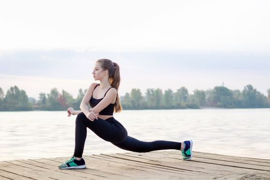 Young pretty slim fitness sporty woman stretches her legs during training workout exercises outdoor at river coast in the morning