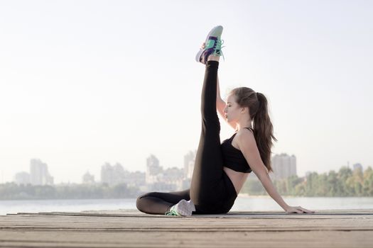 Young pretty slim fitness sporty woman stretches her legs during training workout exercises outdoor at river coast in the morning