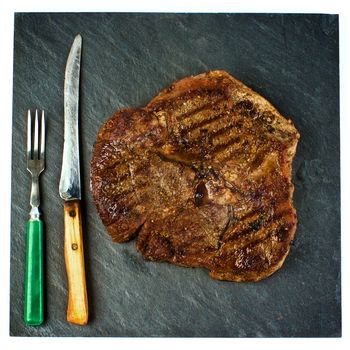 Delicious Grilled Steak Beef Shank on Black Stone Plate with Retro Fork and Knife Cross Section on White background