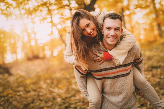 Beautiful young couple enjoying a piggyback in sunny forest in autumn colors. Looking at camera.