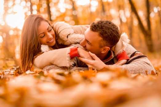 Beautiful smiling couple enjoying in sunny forest in autumn colors. They are lying on the falls leaves and having fun.