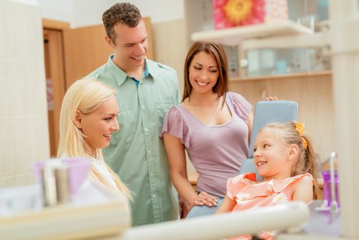 Happy family at visit in the dentist office. Female dentist talking with little girl, her parents standing next to her.