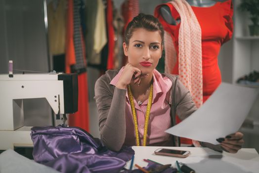 Vintage toned photo of a young woman designer sits in front of the sewing machine, holding sketches of clothing and looking at camera.