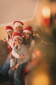 Beautiful smiling family are gathered together by a Christmas tree at the home. Looking at camera and wearing Santa hats. 