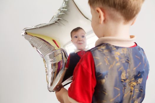 Boy grimacing and playing the ape with star-shaped balloons in studio. Kid looks and rejoices at his reflection in foil balloon. Child laughing looking at the reflection in a distorted mirror