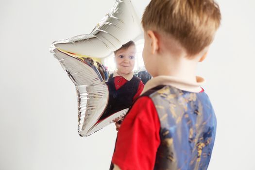 Boy grimacing and playing the ape with star-shaped balloons in studio. Kid looks and rejoices at his reflection in foil balloon. Child drew a smiley face