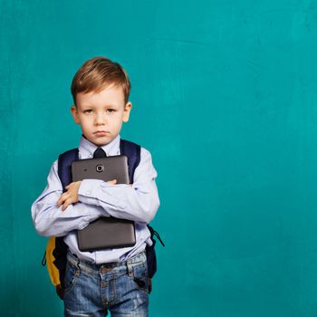 Cheerful little boy with big backpack holding digital tablet against blue background. Looking at camera. School concept. Back to School