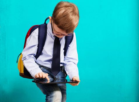 Cheerful little boy with big backpack holding digital tablet against blue background. Child playing games on a touch pad. School concept. Back to School