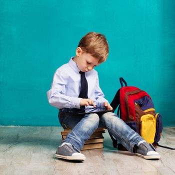 Cheerful little boy with big backpack holding digital tablet against blue background. Child playing games on a touch pad. School concept. Back to School