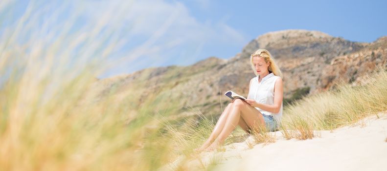 Relaxed woman enjoying sun, freedom and good book an beautiful sandy beach of Balos in Greece. Young lady reading, feeling free and relaxed. Vacations, freedom, happiness, enjoyment and well being.