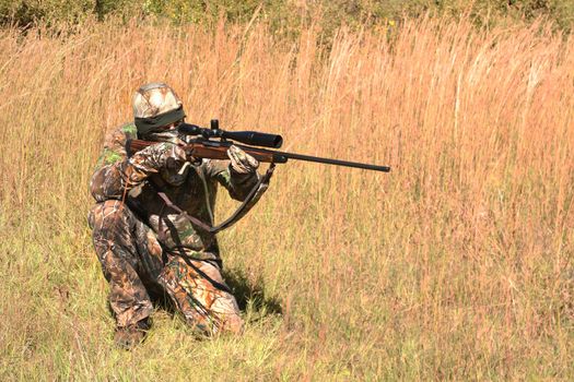 Hunter in grassy field with sniper rifle looking through scope at target