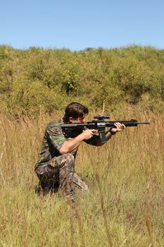 Camouflaged hunter in grassy area with sniper rifle looking through scope at target.