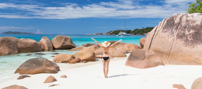 Woman arms rised, wearing black bikini and beach hat, enjoying amazing view on Anse Lazio beach on Praslin Island, Seychelles. Summer vacations on picture perfect tropical beach concept.