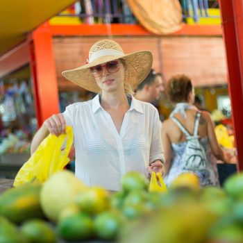 Female traveler wearing elegant colonial style white tunic and hat buying fresh tropical fruit on traditional Victoria food market on Seychelles islands.