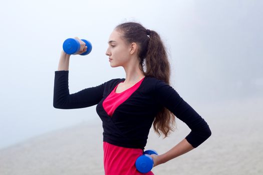 Young pretty slim fitness sporty woman exercises with weights dumbells during training workout outdoor