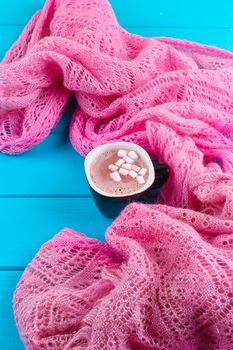 Cozy winter home background, cup of hot cocoa with marshmallow, old vintage books and warm knitted sweater on white painted wooden board background.