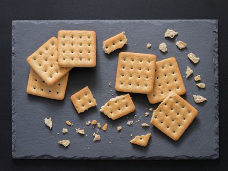 Square crackers with pieces and crumbs on slate gray background. Dry salt cracker cookies with fiber and dry spices. Top view or flat lay.