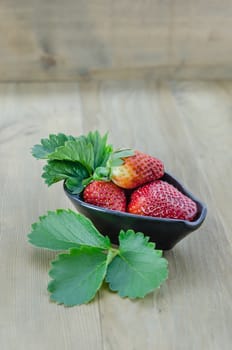 fresh strawberry fruits in black bowl with green leaves over wooden background