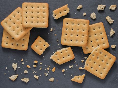 Square crackers with pieces and crumbs on slate gray background. Dry salt cracker cookies with fiber and dry spices. Top view or flat lay.