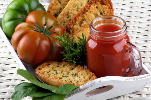 Fresh Tomato Juice in Glass Jar with Raw Tomatoes, Crispy Bread and Fresh Herbs into White Wooden Tray closeup on Wicker background