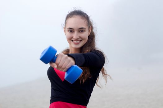 Young pretty slim fitness sporty woman exercises with weights dumbells during training workout outdoor