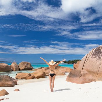 Woman arms rised, wearing black bikini and beach hat, enjoying amazing view on Anse Lazio beach on Praslin Island, Seychelles. Summer vacations on picture perfect tropical beach concept.