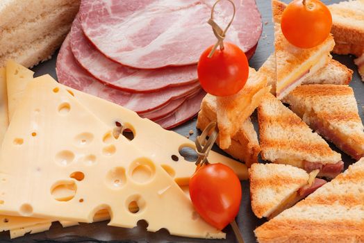 Slices of cheese, ham, bread and marinated cherry tomatoes sandwiches on a slate board