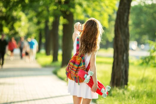 Portrait of lovely urban girl in white dress with a pink skateboard. Happy smiling woman. Girl holding a plastic skate board outdoors. City life. Back view