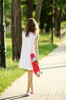 Portrait of lovely urban girl in white dress with a pink skateboard. Happy smiling woman. Girl holding a plastic skate board outdoors. City life. Back view