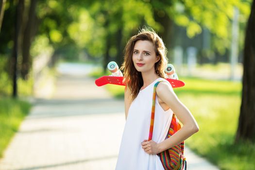 Portrait of lovely urban girl in white dress with a pink skateboard. Happy smiling woman. Girl holding a plastic skate board outdoors. City life.