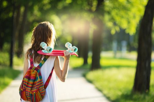 Portrait of lovely urban girl in white dress with a pink skateboard. Happy smiling woman. Girl holding a plastic skate board outdoors. City life. Back view