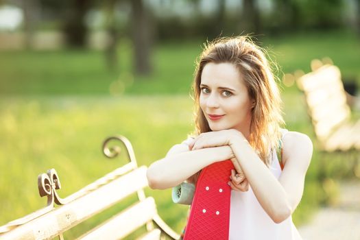 Lovely urban girl sitting on a bench in a city park. Portrait of a happy smiling young woman with a pink skateboard.